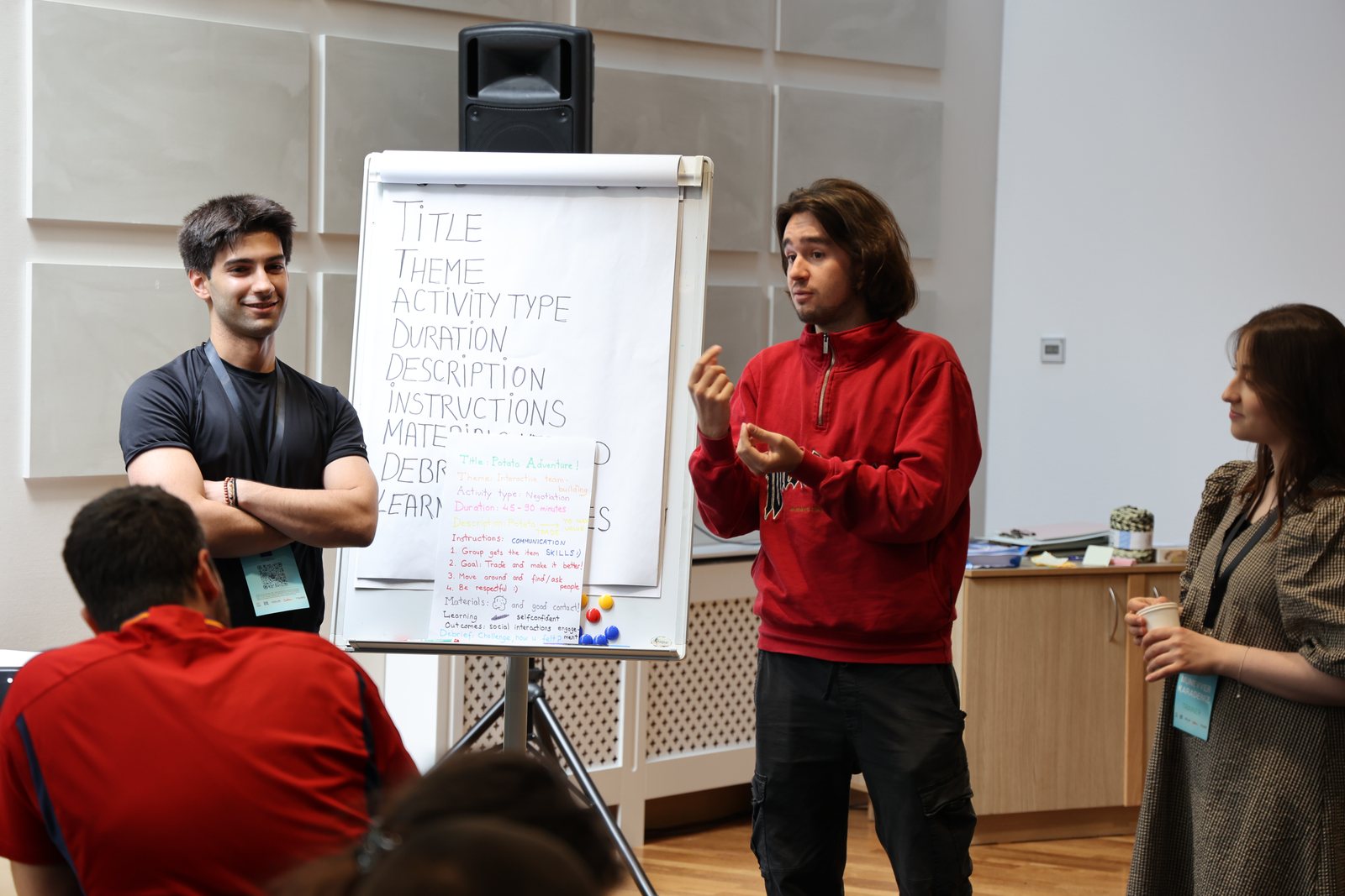 Three participants present their own activity next to a flipchart listing title, theme, activity type, duration, description and instructions