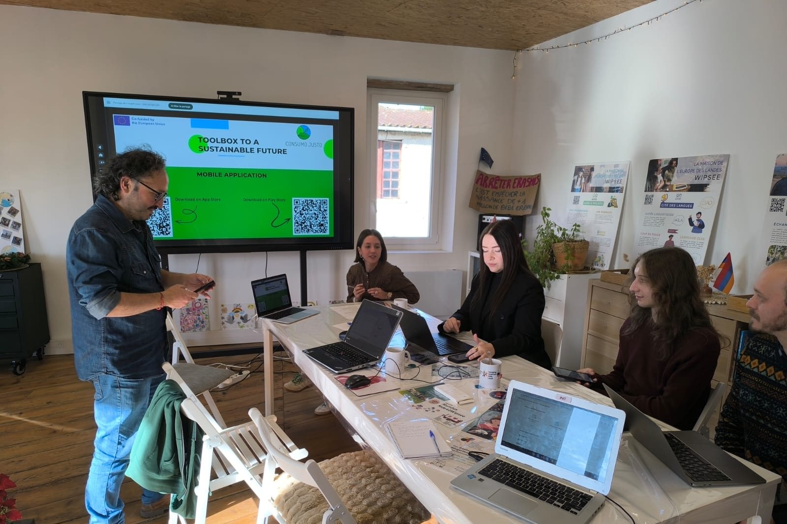 A partner presents the app on a large screen showing two QR codes while colleagues follow on their laptops