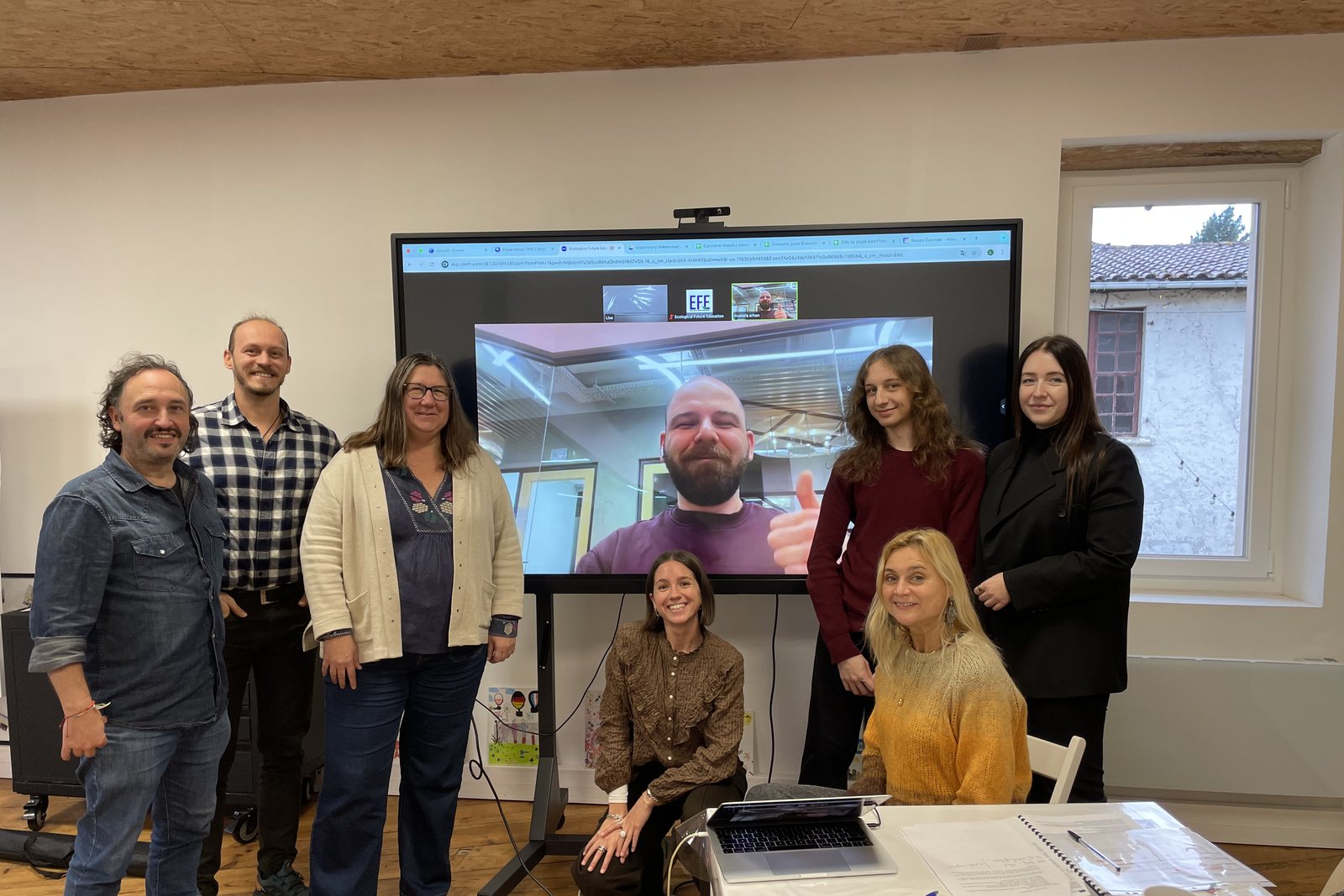 People from the partner organisations stand in front of a screen on which a colleague joins by video call