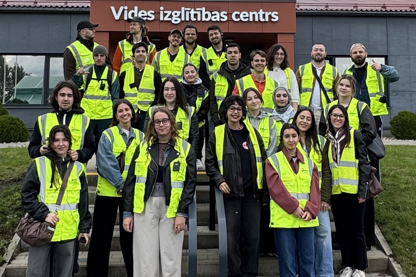 Participants in yellow safety vests stand on the steps of the environmental education centre at Getliņi