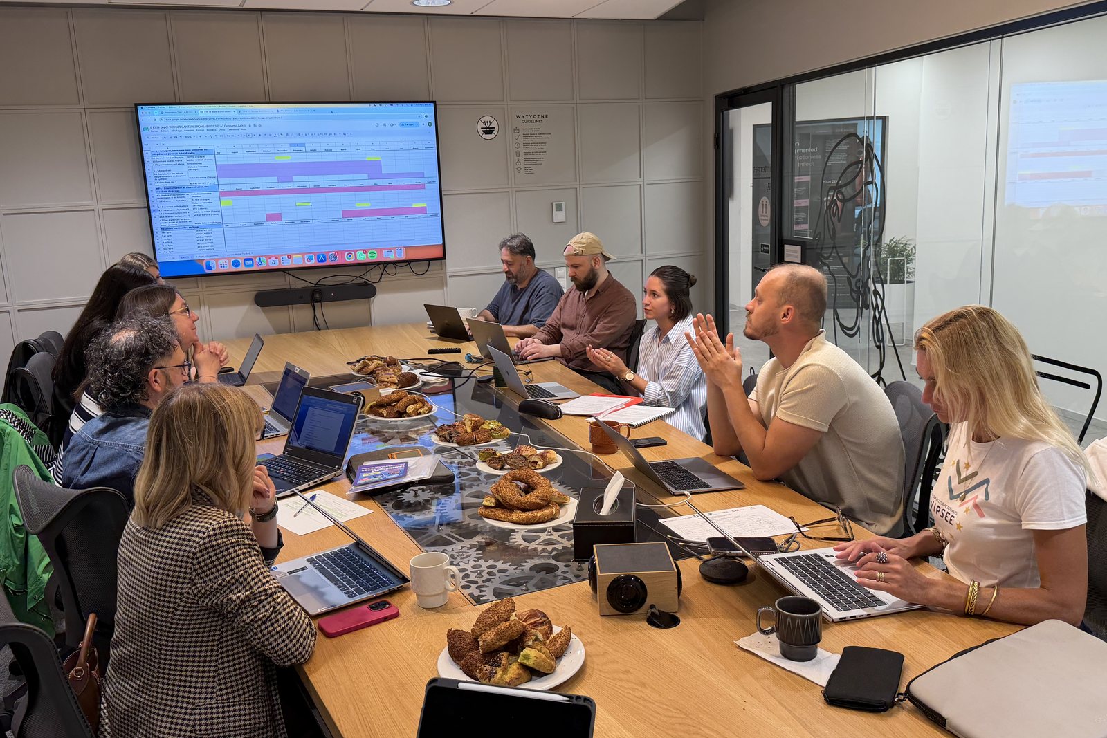 Partners sit around a long meeting table with laptops while a planning chart is shown on a wall screen
